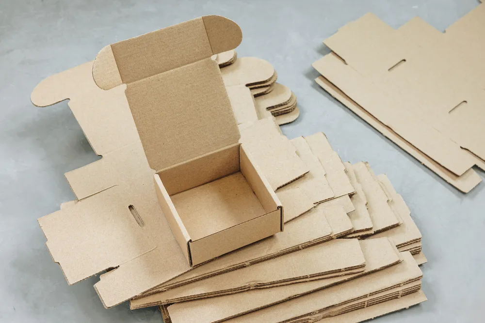 Persona preparando un envío, doblando las solapas de una de las varias cajas y empaques de cartón en una mesa.